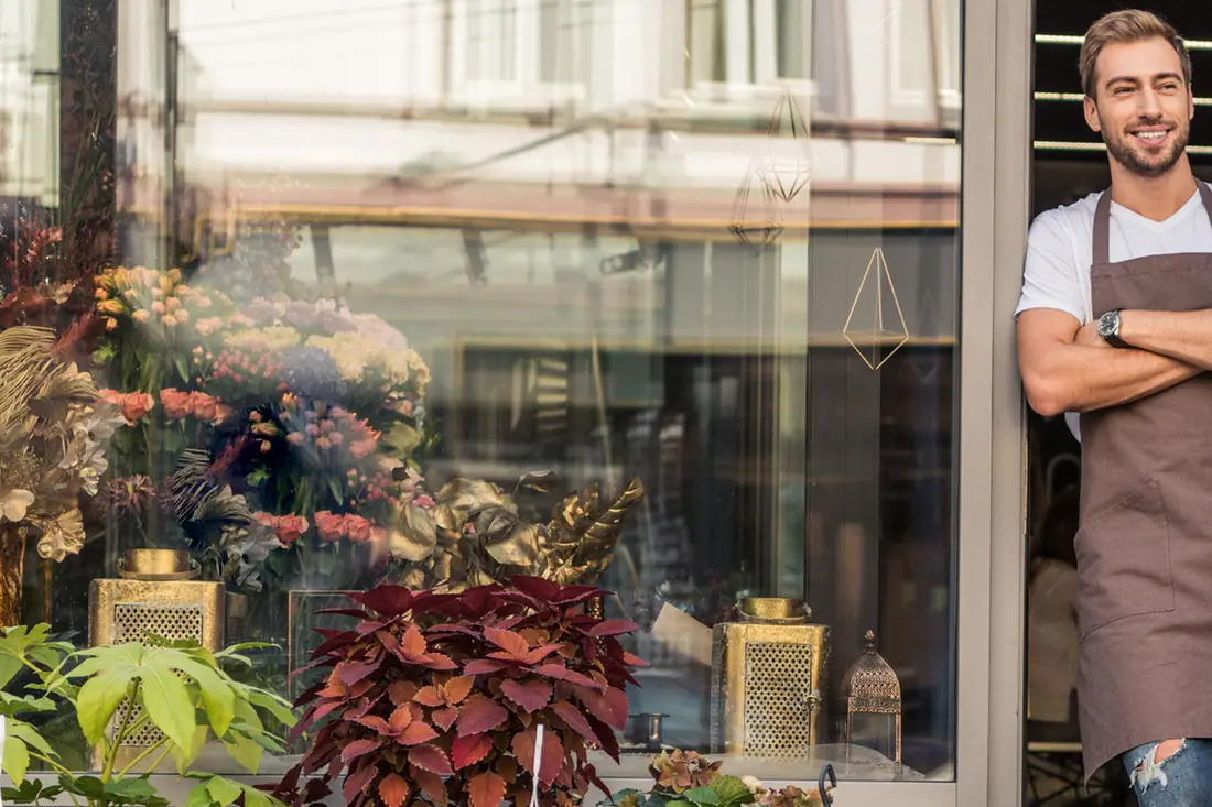 A florist in an apron, arms folded, in the doorway of the shop he runs.