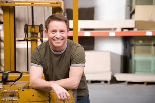 A warehouse owner leaning on his forklift, smiling, stock racked behind him.